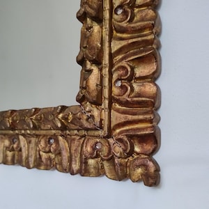Könnte beinhalten: Nahaufnahme eines Abschnitts eines goldfarbenen, verzierten Bilderrahmens. Der Rahmen hat ein detailliertes, geschnitztes Design mit floralen und scrollartigen Motiven. Die Oberfläche des Rahmens hat ein antikes Aussehen mit Variationen im Goldton.
