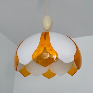Peut inclure: Un luminaire suspendu blanc et orange. Le luminaire a une forme de fleur avec des p&eacute;tales blancs et des accents orange. Il est suspendu au plafond par un fin cordon blanc cass&eacute;.