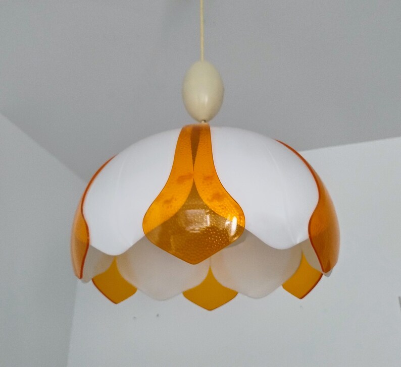 Peut inclure: Un luminaire suspendu blanc et orange. Le luminaire a une forme de fleur avec des p&eacute;tales blancs et des accents orange. Il est suspendu au plafond par un fin cordon blanc cass&eacute;.