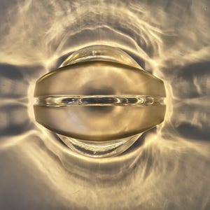 Könnte beinhalten: Ein klares Glasobjekt mit abgerundeter, symmetrischer Form und einem horizontalen Band in der Mitte. Das Glas strahlt ein warmes, goldenes Licht aus, das komplexe, fließende und wellenartige Muster auf die umgebende Oberfläche wirft.