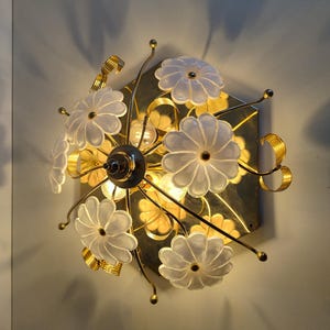 May include: A wall-mounted light fixture with a floral design. It features white, flower-shaped glass shades, gold-coloured metal accents, and a central light source. The fixture is mounted on a gold-toned, hexagonal base.