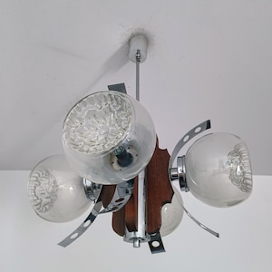 Könnte beinhalten: Eine Deckenleuchte aus Chrom und Holz mit vier klaren Glaskugeln als Schirmen. Die Schirme haben eine strukturierte Oberfläche und hängen an einer zentralen Chromstange.