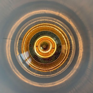 Pode incluir: Close-up de uma lâmpada Edison acesa. A lâmpada tem um exterior de vidro transparente com um filamento dourado e componentes internos. A luz projeta um brilho quente, com anéis concêntricos de luz irradiando para fora contra um fundo azul pálido.