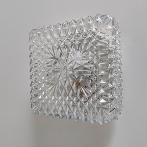 Könnte beinhalten: Eine klare, quadratische Glasschale mit einem strukturierten, geometrischen Muster. Die Schale hat ein facettiertes Design, das einen funkelnden Effekt erzeugt. Die Kanten sind leicht uneben, was ihren einzigartigen Charakter unterstreicht. Dieses dekorative Stück könnte zum Servieren oder Ausstellen verwendet werden.