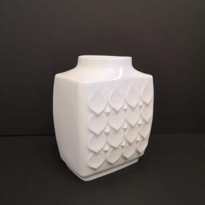 May include: A white ceramic vase with a square base and a slightly flared neck. The vase has a repeating pattern of raised, leaf-shaped designs on its sides. The background is a solid black.