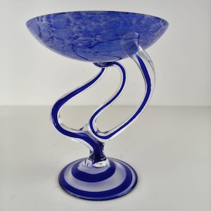 May include: A decorative glass bowl featuring a deep blue mottled pattern. The bowl is supported by a clear glass stem with swirling blue accents that curves upward to meet the base of the bowl, resting on a circular base with blue stripes.
