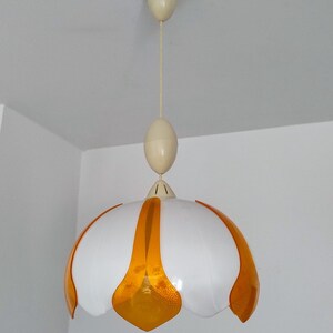 Peut inclure: Un luminaire suspendu blanc et orange. La lampe a un abat-jour blanc en forme de fleur avec des accents orange. Le luminaire est suspendu au plafond par un cordon cr&egrave;me et une pi&egrave;ce ovale assortie.