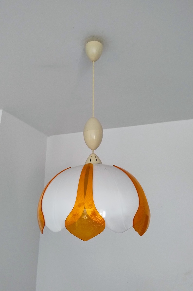 Peut inclure: Un luminaire suspendu blanc et orange. La lampe a un abat-jour blanc en forme de fleur avec des accents orange. Le luminaire est suspendu au plafond par un cordon cr&egrave;me et une pi&egrave;ce ovale assortie.