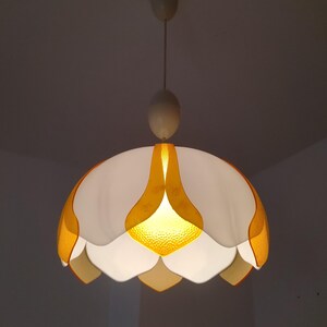 Peut inclure: Suspension luminaire vintage en forme de fleur. La lampe pr&eacute;sente une palette de couleurs blanche et jaune, avec un centre jaune textur&eacute; et des abat-jours en forme de p&eacute;tales. Le luminaire est suspendu au plafond par un cordon.