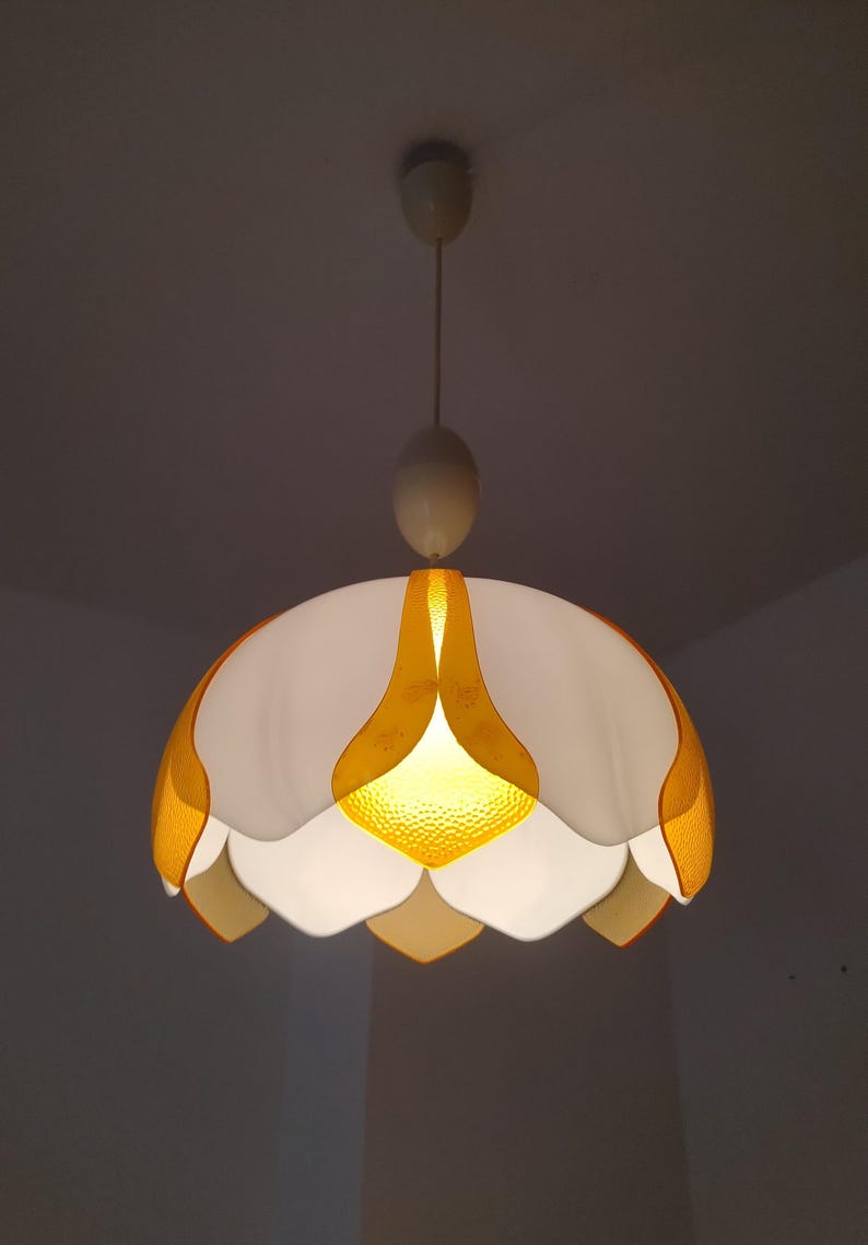 Peut inclure: Suspension luminaire vintage en forme de fleur. La lampe pr&eacute;sente une palette de couleurs blanche et jaune, avec un centre jaune textur&eacute; et des abat-jours en forme de p&eacute;tales. Le luminaire est suspendu au plafond par un cordon.