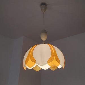 Peut inclure: Un luminaire suspendu blanc et orange. Le luminaire a une forme arrondie en forme de fleur avec des accents en forme de p&eacute;tales orange. Il est suspendu &agrave; un cordon blanc et &agrave; un support de plafond, offrant une lueur chaleureuse.