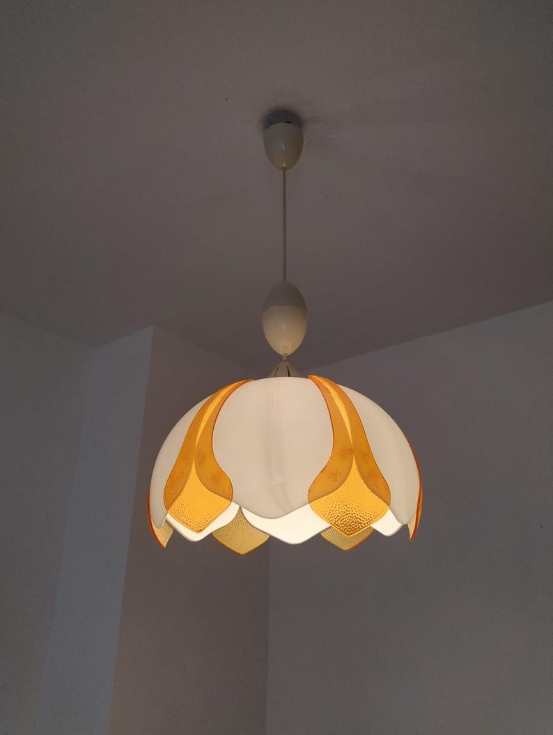 Peut inclure: Un luminaire suspendu blanc et orange. Le luminaire a une forme arrondie en forme de fleur avec des accents en forme de p&eacute;tales orange. Il est suspendu &agrave; un cordon blanc et &agrave; un support de plafond, offrant une lueur chaleureuse.