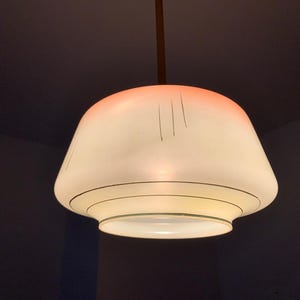 May include: A vintage pendant light fixture with a white glass shade. The shade has a rounded, bell-like shape with a reddish-orange gradient at the top. Black lines and circles decorate the shade. The fixture hangs from a brown rod.