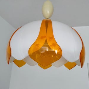 Peut inclure: Suspension luminaire blanc et orange. Le luminaire a une forme de fleur avec des p&eacute;tales blancs et des accents orange. Il est suspendu au plafond par un cordon blanc et une pi&egrave;ce ovale cr&egrave;me.