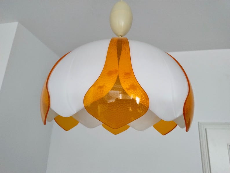 Peut inclure: Suspension luminaire blanc et orange. Le luminaire a une forme de fleur avec des p&eacute;tales blancs et des accents orange. Il est suspendu au plafond par un cordon blanc et une pi&egrave;ce ovale cr&egrave;me.
