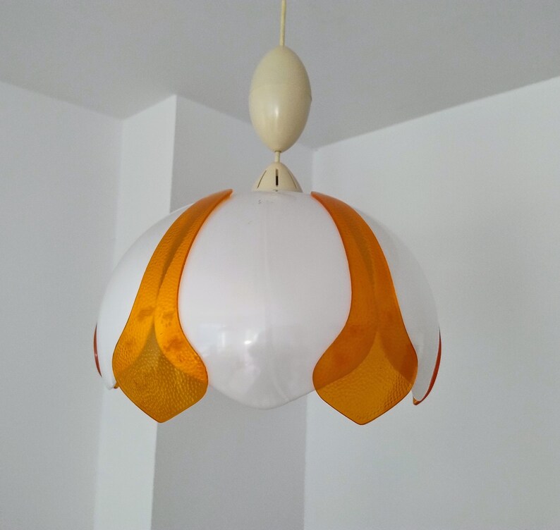 Peut inclure: Un luminaire suspendu vintage avec un abat-jour blanc en forme de fleur et des accents orange. La lumi&egrave;re est suspendue &agrave; un luminaire ovale de couleur cr&egrave;me. Le luminaire est suspendu &agrave; un cordon fin de couleur cr&egrave;me.