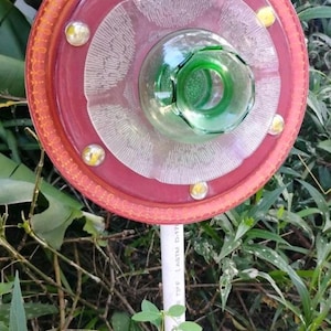 May include: A handcrafted garden art piece resembling a flower. It features a circular design with a red outer rim, clear glass elements, and a green glass bottle center. The piece is adorned with yellow accents and mounted on a white pole.
