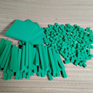 May include: Green math manipulatives, including squares, rods, and small cubes, are arranged on a wooden surface. The squares have a grid pattern, and the rods are rectangular. The cubes are small and numerous.