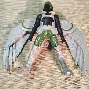 Pu&ograve; includere: Action figure di Vulture con ali articolate. La figura presenta un elmetto verde e nero, un busto nero e bianco e gambe verdi e bianche. Le ali sono grigie con dettagli verdi. La figura &egrave; in posa con le gambe divaricate.