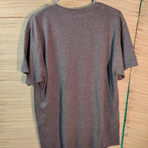 Puede incluir: Una camiseta gris jaspeada colgada de una percha de pl&aacute;stico gris. La camiseta de manga corta est&aacute; hecha de un material suave y se muestra sobre un fondo marr&oacute;n claro.