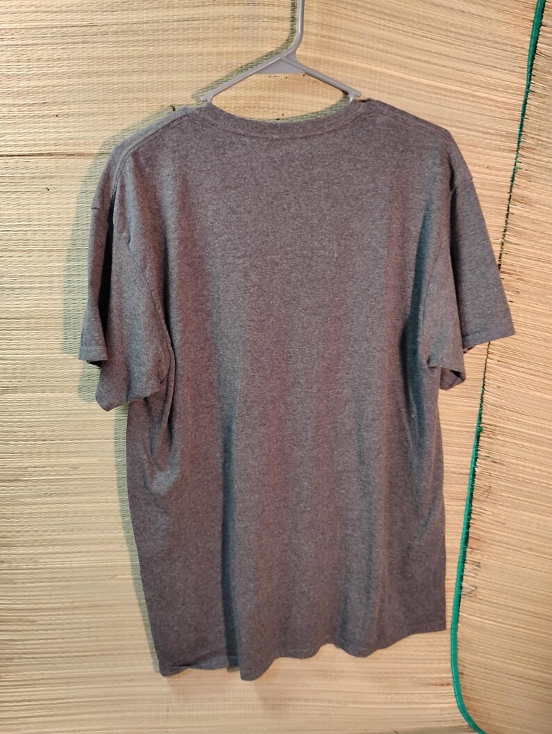 Puede incluir: Una camiseta gris jaspeada colgada de una percha de pl&aacute;stico gris. La camiseta de manga corta est&aacute; hecha de un material suave y se muestra sobre un fondo marr&oacute;n claro.