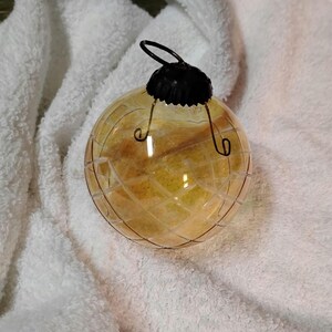 May include: A clear, round Christmas ornament with a yellow interior and a black metal cap. The ornament has a grid-like pattern etched on its surface and is suspended by a wire loop. The ornament is resting on a white, textured surface.