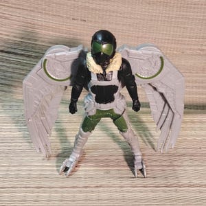Pu&ograve; includere: Una action figure di Vulture, con un casco nero e visiera verde, una tuta nera e verde e grandi ali grigie con dettagli verdi. La figura ha un colletto di pelliccia marrone e un'armatura per le gambe dettagliata.