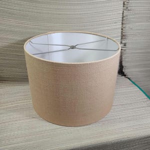 May include: A cylindrical lamp shade featuring a textured, light beige fabric exterior and a smooth white interior lining. The top opening contains a silver-toned metal spider fitter with four spokes meeting at a central ring for attachment.