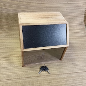 May include: A wooden donation box featuring a light brown natural wood frame, a rectangular black chalkboard panel on the front, and a clear acrylic viewing window below. A horizontal slot is located on the top surface, and two metal keys are included.