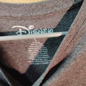 Puede incluir: Una camiseta gris oscuro con el logo de Disney en blanco. La etiqueta muestra la composici&oacute;n de la tela, las instrucciones de lavado y la talla es L. La camiseta est&aacute; hecha en Honduras.