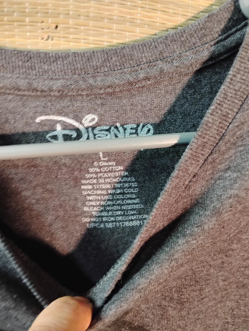 Puede incluir: Una camiseta gris oscuro con el logo de Disney en blanco. La etiqueta muestra la composici&oacute;n de la tela, las instrucciones de lavado y la talla es L. La camiseta est&aacute; hecha en Honduras.