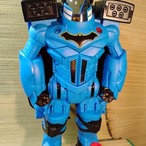 May include: A blue Batman action figure with a black bat symbol on the chest. The toy has a clear helmet, black accents, and missile launchers on the shoulders. The figure is standing on a patterned surface.