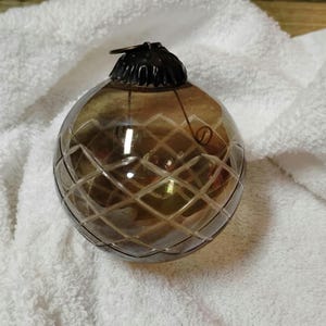 May include: A brown glass Christmas ornament with a diamond pattern. The ornament has a black metal cap and a loop for hanging. The ornament is sitting on a white, textured surface.