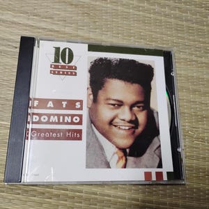 May include: A Fats Domino "Greatest Hits" CD in a clear plastic case. The album cover features a portrait of Fats Domino and the text "10 Best Series". The CD case has a black spine.