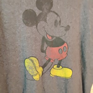 Puede incluir: Una camiseta gris con un gr&aacute;fico desgastado de Mickey Mouse. El personaje de dibujos animados est&aacute; representado en negro, rojo y amarillo, sonriendo con las manos a la espalda. La camiseta tiene un aspecto vintage y desgastado.