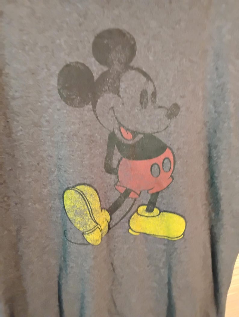 Puede incluir: Una camiseta gris con un gr&aacute;fico desgastado de Mickey Mouse. El personaje de dibujos animados est&aacute; representado en negro, rojo y amarillo, sonriendo con las manos a la espalda. La camiseta tiene un aspecto vintage y desgastado.