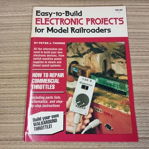May include: A softcover book titled Easy-to-Build ELECTRONIC PROJECTS for Model Railroaders by Peter J. Thorne. The cover features a red and white design with text, a model train scene, and a handheld throttle controller with speed and direction labels.
