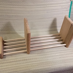 May include: A light-colored wooden rack featuring three vertical panels connected by a series of horizontal dowels. The structure is divided into two sections by a central upright panel, with two outer panels providing support at each end of the frame.