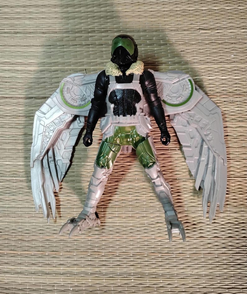 Pu&ograve; includere: Action figure di Vulture con ali articolate. La figura presenta un casco nero e verde, una tuta nera e un'armatura argentata e verde. Le ali sono dettagliate con un design a forma di piuma. La figura &egrave; posata su una superficie tessuta.