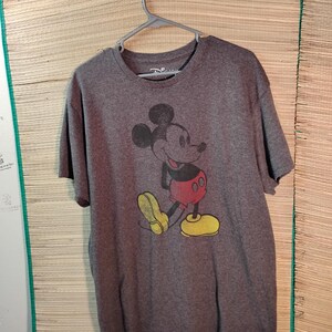 Puede incluir: Una camiseta gris con un gr&aacute;fico desgastado de Mickey Mouse. El personaje de dibujos animados se representa en su pose cl&aacute;sica, con pantalones cortos rojos, zapatos amarillos y guantes negros. La camiseta est&aacute; colgada de una percha de pl&aacute;stico gris.