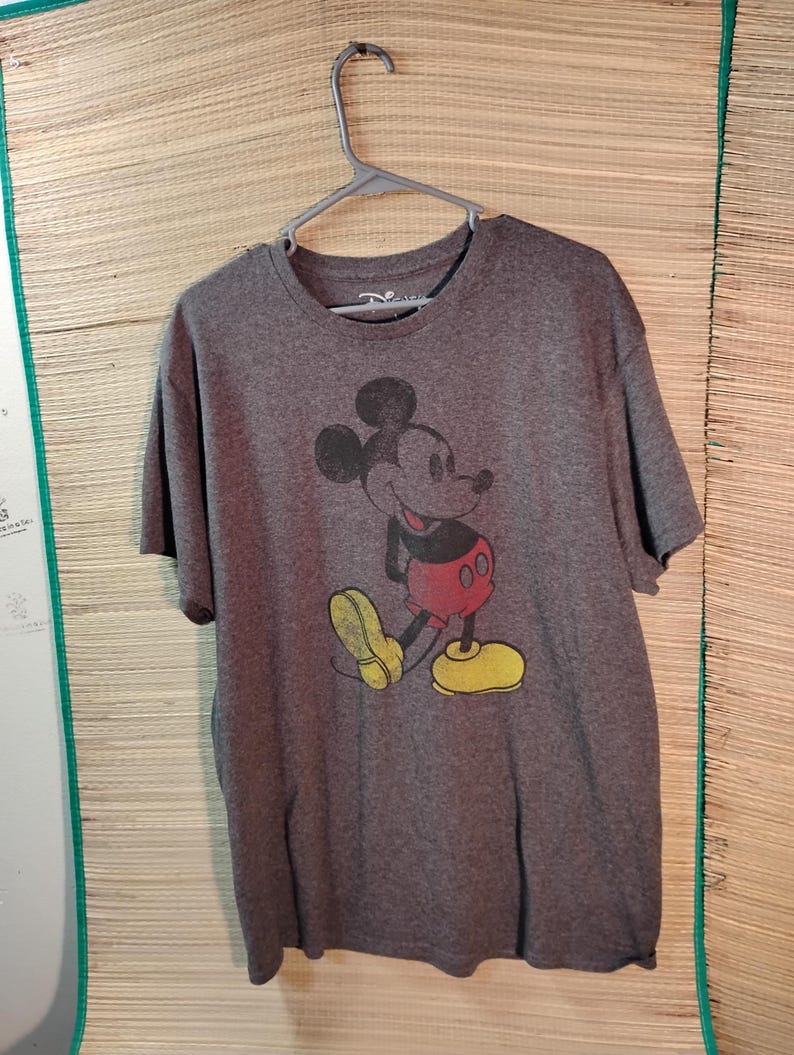 Puede incluir: Una camiseta gris con un gr&aacute;fico desgastado de Mickey Mouse. El personaje de dibujos animados se representa en su pose cl&aacute;sica, con pantalones cortos rojos, zapatos amarillos y guantes negros. La camiseta est&aacute; colgada de una percha de pl&aacute;stico gris.