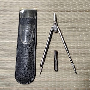 May include: A silver drafting compass with a needle point and pencil holder, plus a small silver pencil lead container. A black leatherette case with a clear plastic window holds the compass. The tools are on a woven mat.