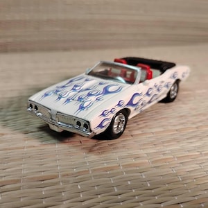 May include: A white convertible toy car with blue flame decals. The car has a black top, red interior, and chrome detailing on the front. The wheels are black.