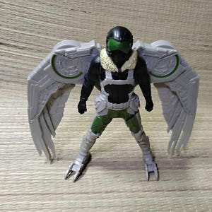 Pu&ograve; includere: Action figure di Vulture, con casco nero e visiera verde, tuta nera e argento, e grandi ali grigie con dettagli verdi. La figura &egrave; in posa con le gambe divaricate.