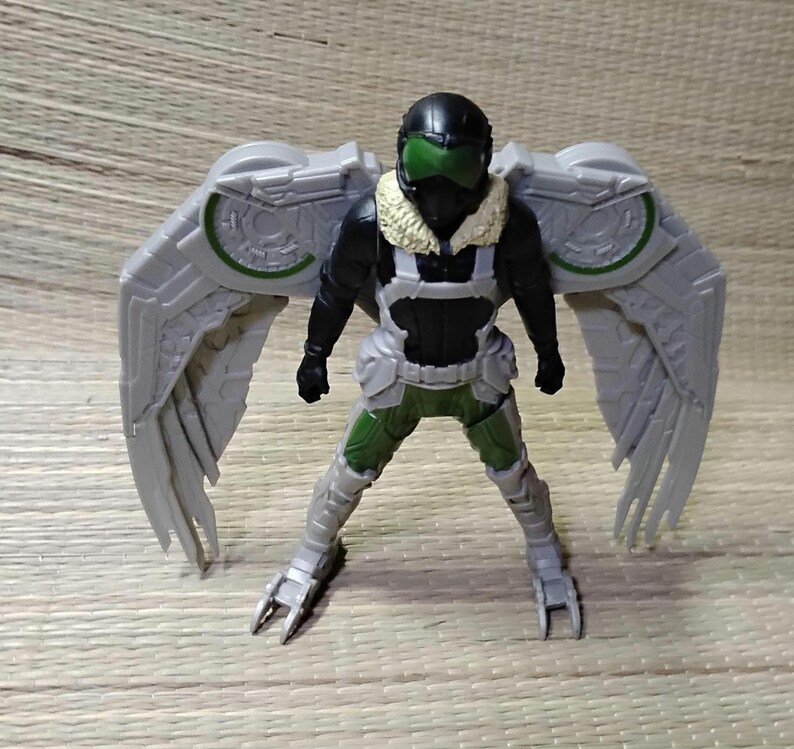 Pu&ograve; includere: Action figure di Vulture, con casco nero e visiera verde, tuta nera e argento, e grandi ali grigie con dettagli verdi. La figura &egrave; in posa con le gambe divaricate.