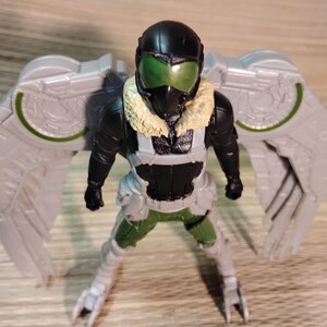 Pu&ograve; includere: Una action figure di Vulture, con un casco nero e visiera verde, una tuta nera e verde e grandi ali grigie. La figura ha un colletto di pelliccia beige ed &egrave; in posa in piedi con le gambe divaricate.
