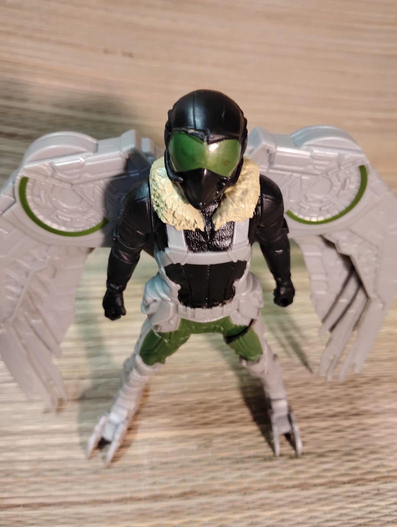 Pu&ograve; includere: Una action figure di Vulture, con un casco nero e visiera verde, una tuta nera e verde e grandi ali grigie. La figura ha un colletto di pelliccia beige ed &egrave; in posa in piedi con le gambe divaricate.