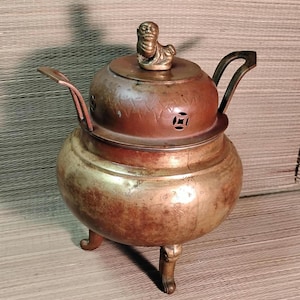 May include: Antique bronze incense burner with a small figurine on the lid. The burner has a round body, three legs, and a handle. The lid has decorative cutouts. The metal has a weathered, aged appearance.