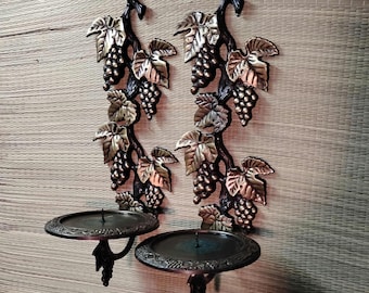 May include: Pair of wall-mounted candle holders with a grape vine design. The metal candle holders feature black stems, bronze-colored leaves, and clusters of grapes. The candle holders have a circular base to hold candles.