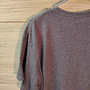 Puede incluir: Una camiseta gris jaspeado cuelga de una percha de pl&aacute;stico gris. La camiseta de manga corta est&aacute; hecha de un material suave. Se ven el cuello y las costuras de los hombros de la camiseta. El fondo es una superficie tejida de color marr&oacute;n claro.